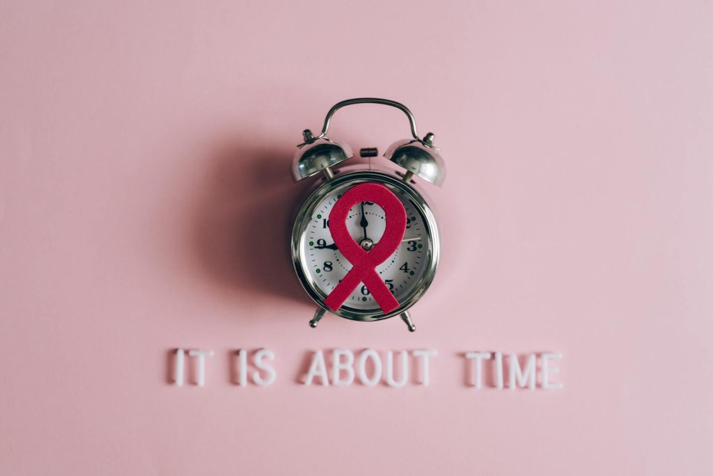 Imagem mostra um relógio analógico com uma fita vermelha representando a luta contra a Aids e a frase "It is about time"